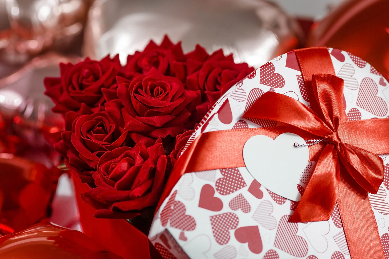 Valentine's day. Red roses in a white box in the form of a heart on the background of foil balls. A gift for women on a holiday. The concept of delivering flowers. floristry and flower shops.